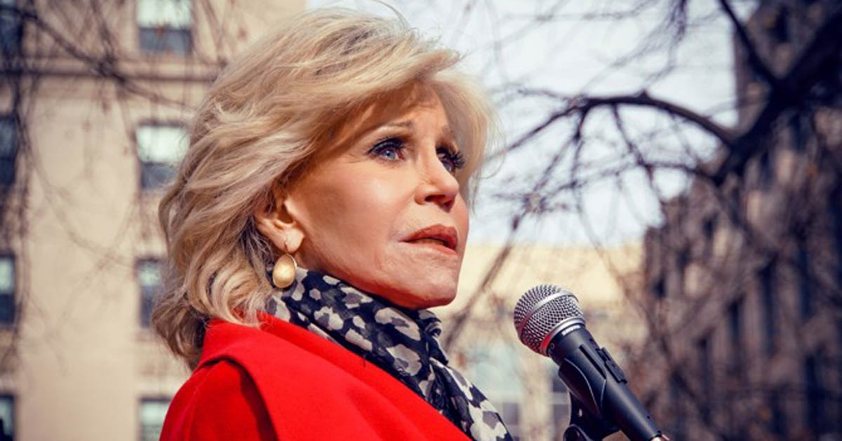 Jane Fonda wears a red coat and speaks into a microphone