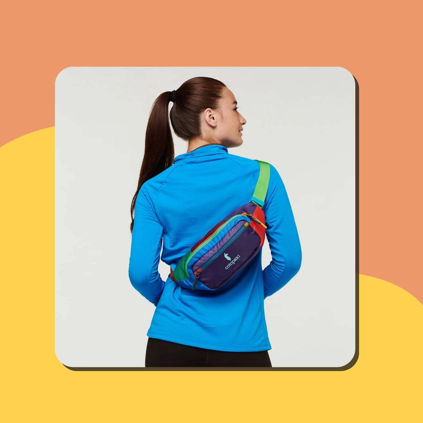 A woman with a brunette ponytail wears a colorful Cotopaxi hip pack