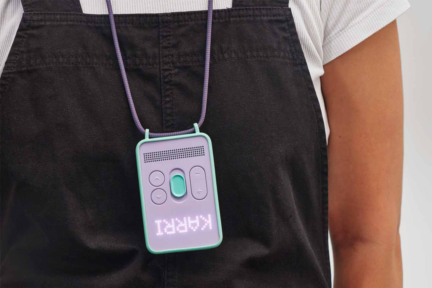A screen-free device sits on a purple lanyard atop black overalls