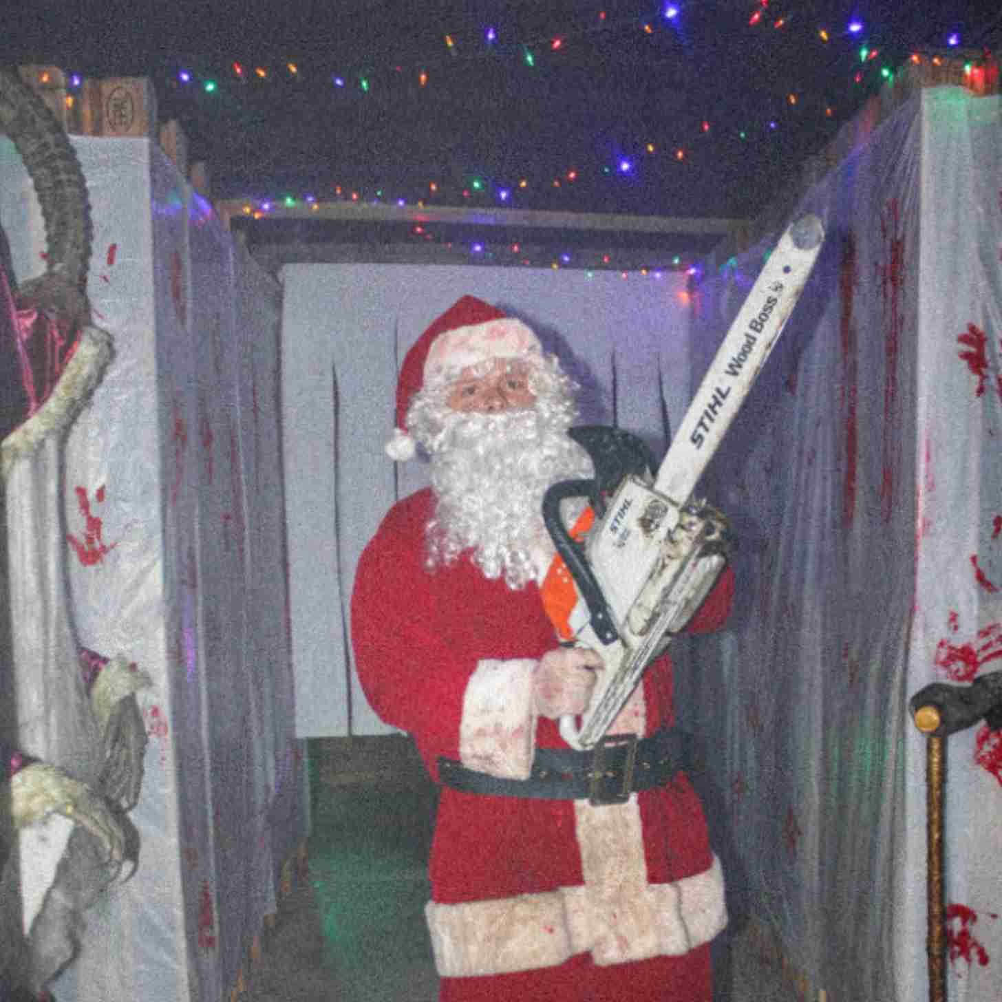 A man in a Santa Claus costume stands in a dark hallway weilding a chainsaw