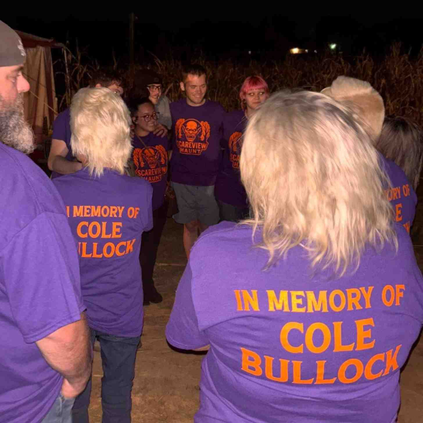 A crowd of people smile and stand close in a cornfield. They are all wearing Scareview Haunt shirts that read "In Memory of Cole Bullock" on the back.