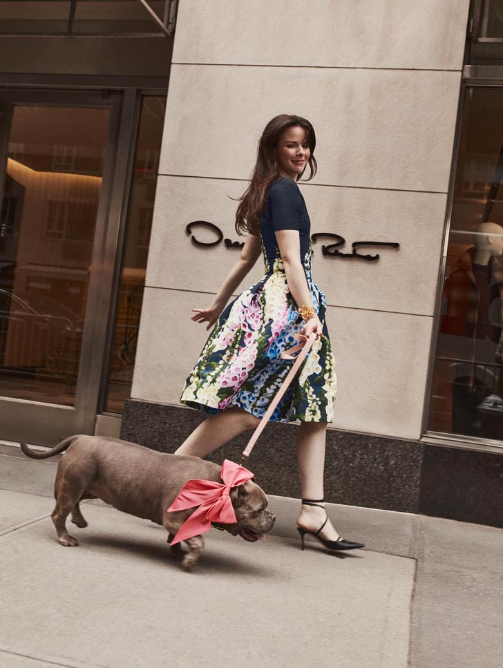 A white woman with long brown hair walks a pit bull in front of Oscar de la Renta's store in New York City. She wears a floral dress and the dog wears a pink bow
