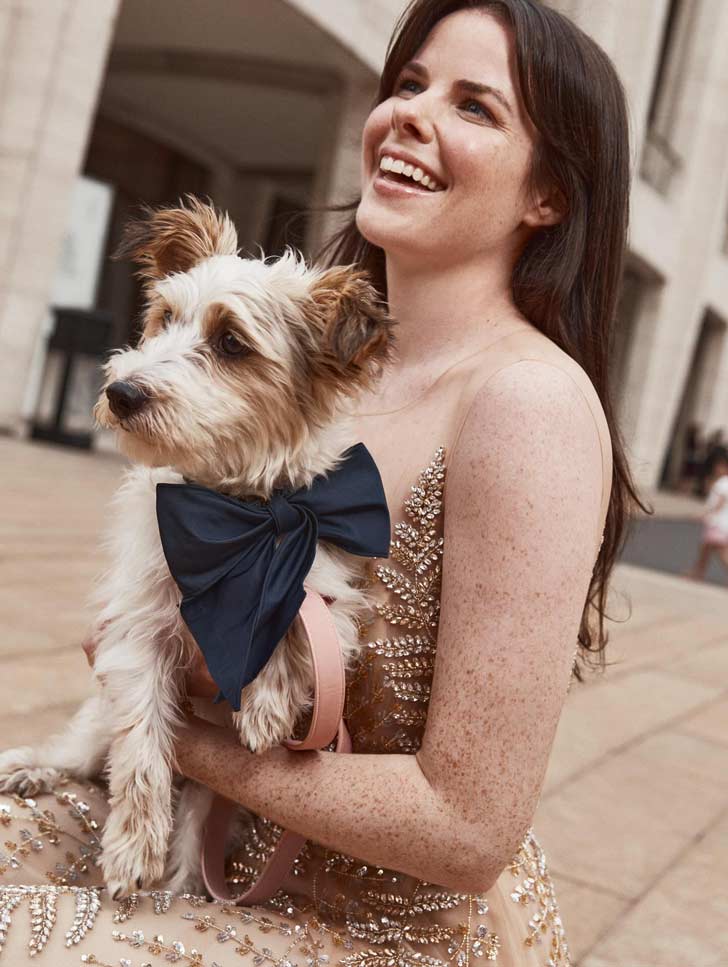 A white woman with brown hair holds a small white terrier, smiling. She is in a gold sequin gown, and the dog is in a navy blue bow
