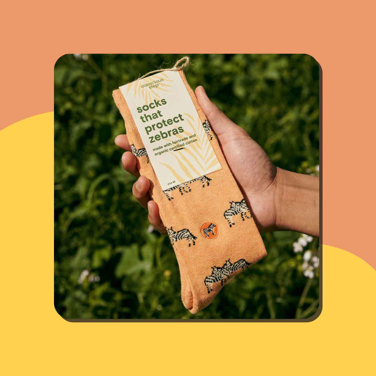 A person holds up a pair of Conscious Step socks that are orange and have zebras on them