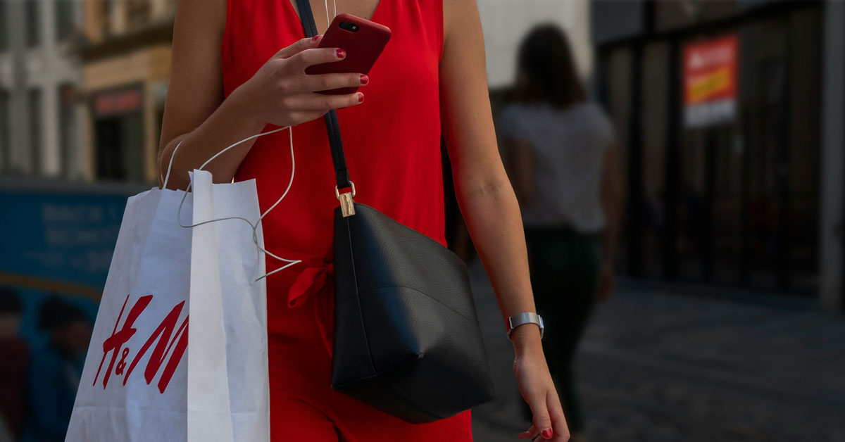 A close-up of a woman's torso, where she carries a white H&M bag on her arm and holds a phone in her hand