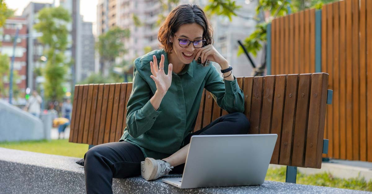 A woman sits on a bench outdoors with a laptop in front of her, taking a video call for work