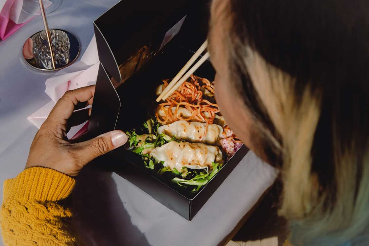 Over a person's shoulder sits a black takeout box filled with dumplings, noodles, and vegetables from Lovebite Dumplings in Phoenix, Arizona