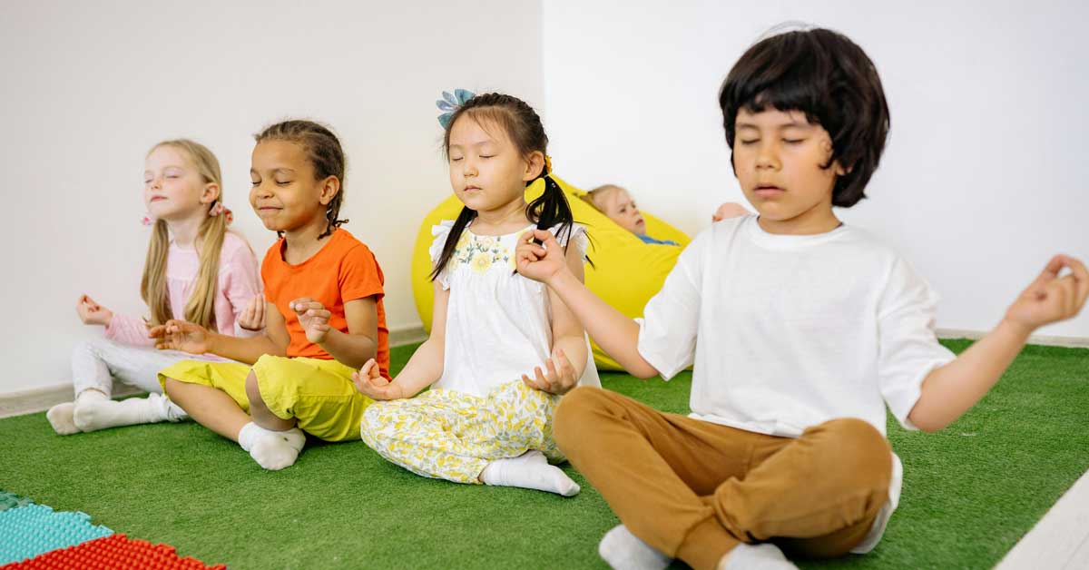 Four racially diverse children meditate on a green mat
