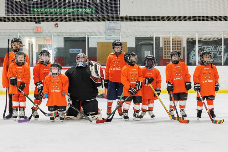 A row of children in orange jerseys stand on the ice with hockey sticks