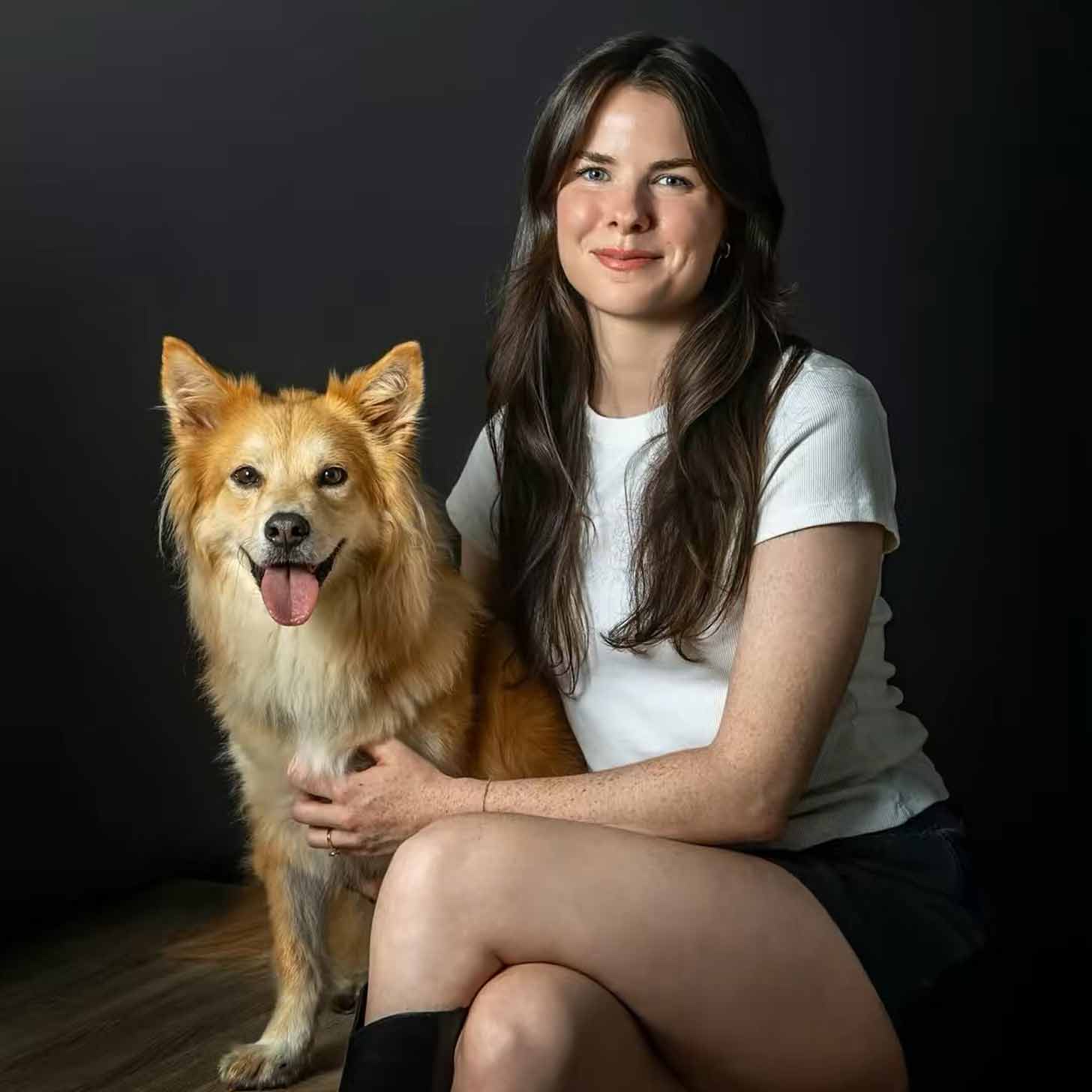 A woman, Isabel Klee, sits next to her dog, a tan fluffy mix with pointy ears, in front of a slate gray backdrop.