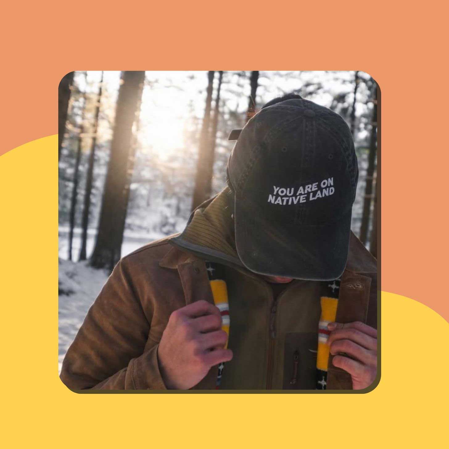 A person wears a black baseball cap that reads "you are on native land"