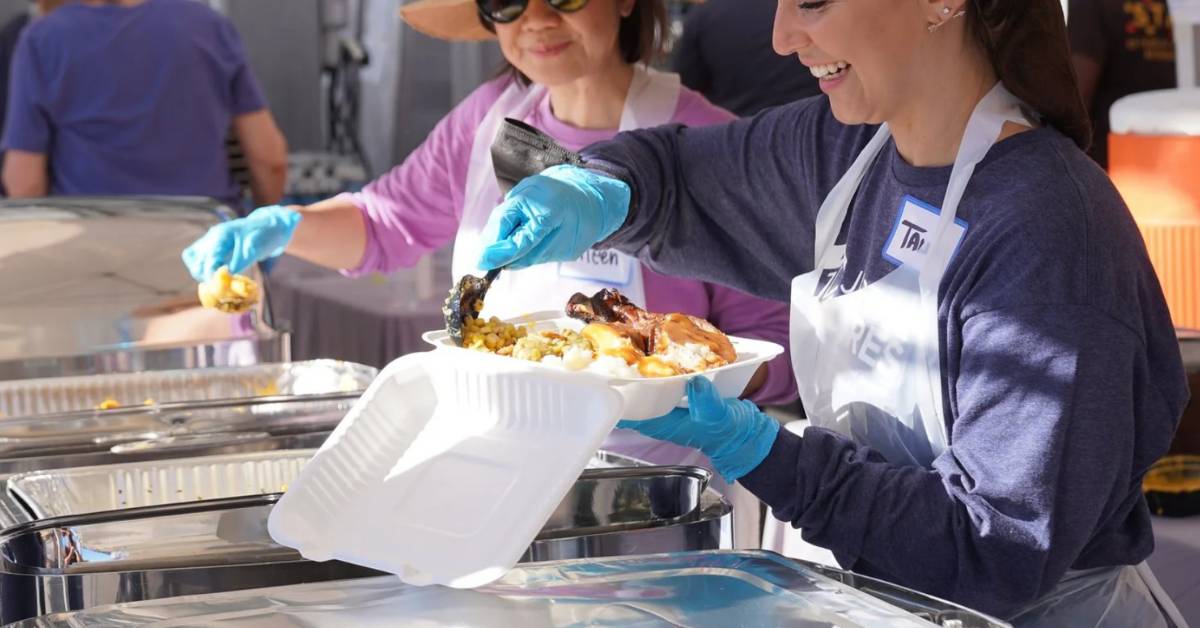 Two women smile as they load food into Styrofoam trays at a food bank