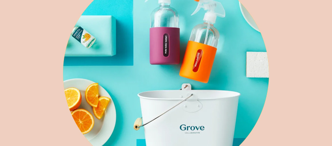 Overhead flatlay of a white cleaning bucket, two glass spray bottles, a plate of oranges and cleaner concentrate bottle on a bright teal background. 