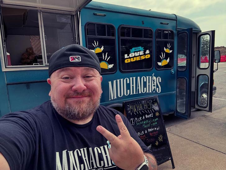 A man with a graying beard and hat throws up a peace sign in front of a blue food truck called Muchachos