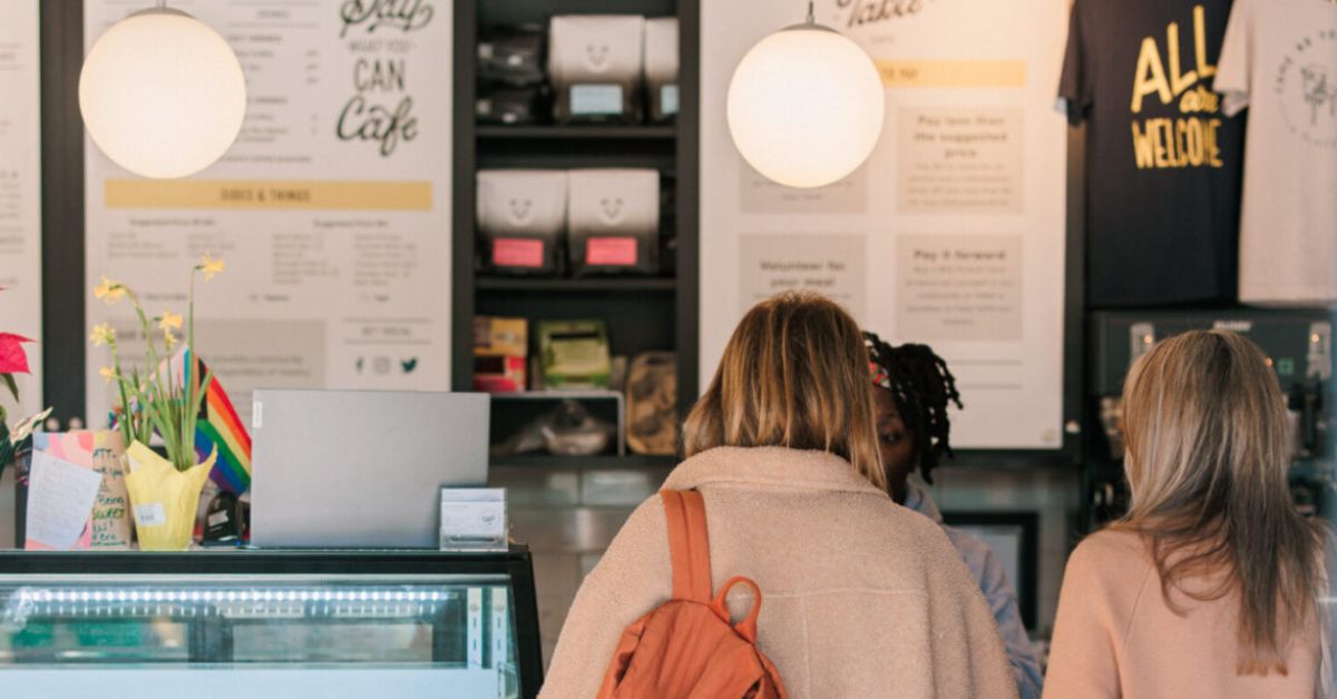 Two women wait in line at a cafe with their backs turned to the camera.