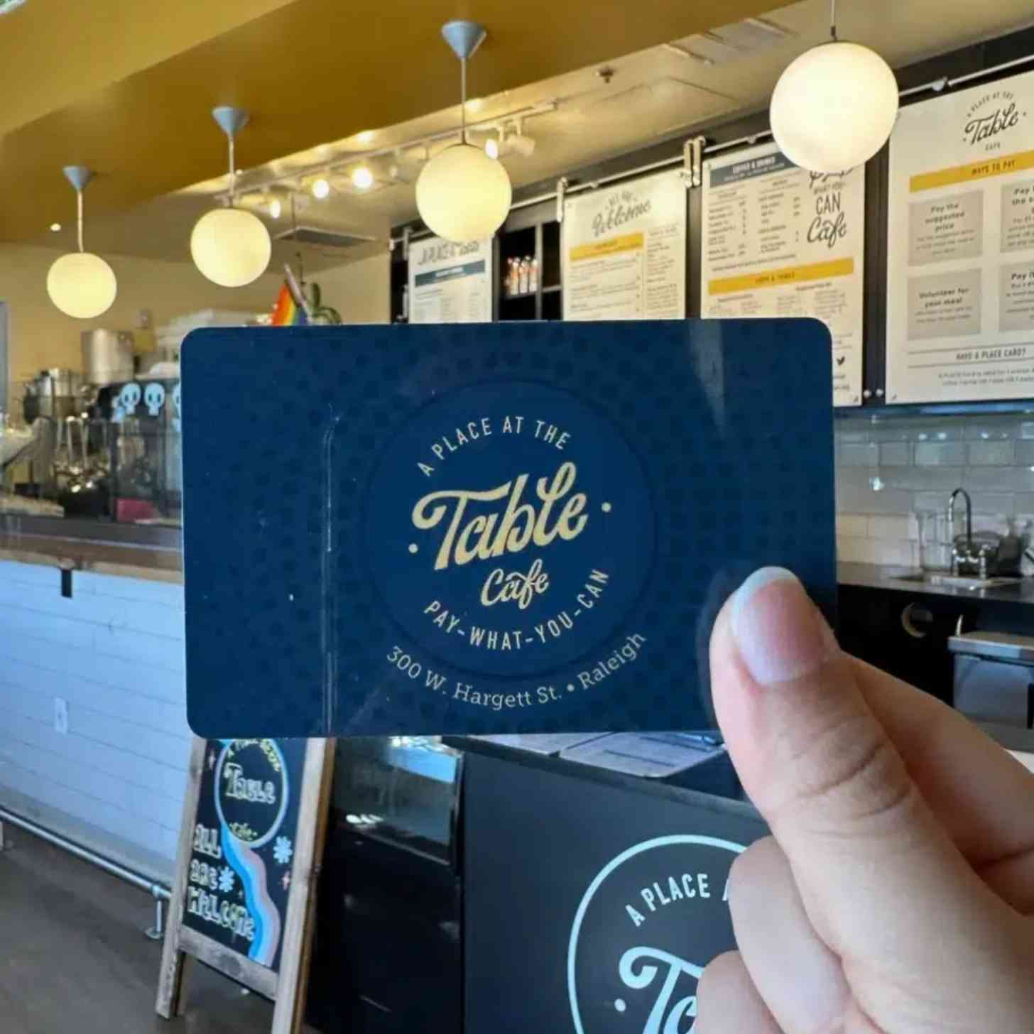 A person holds up a business card that says "A Place At The Table" as they are in the lobby of the very same cafe