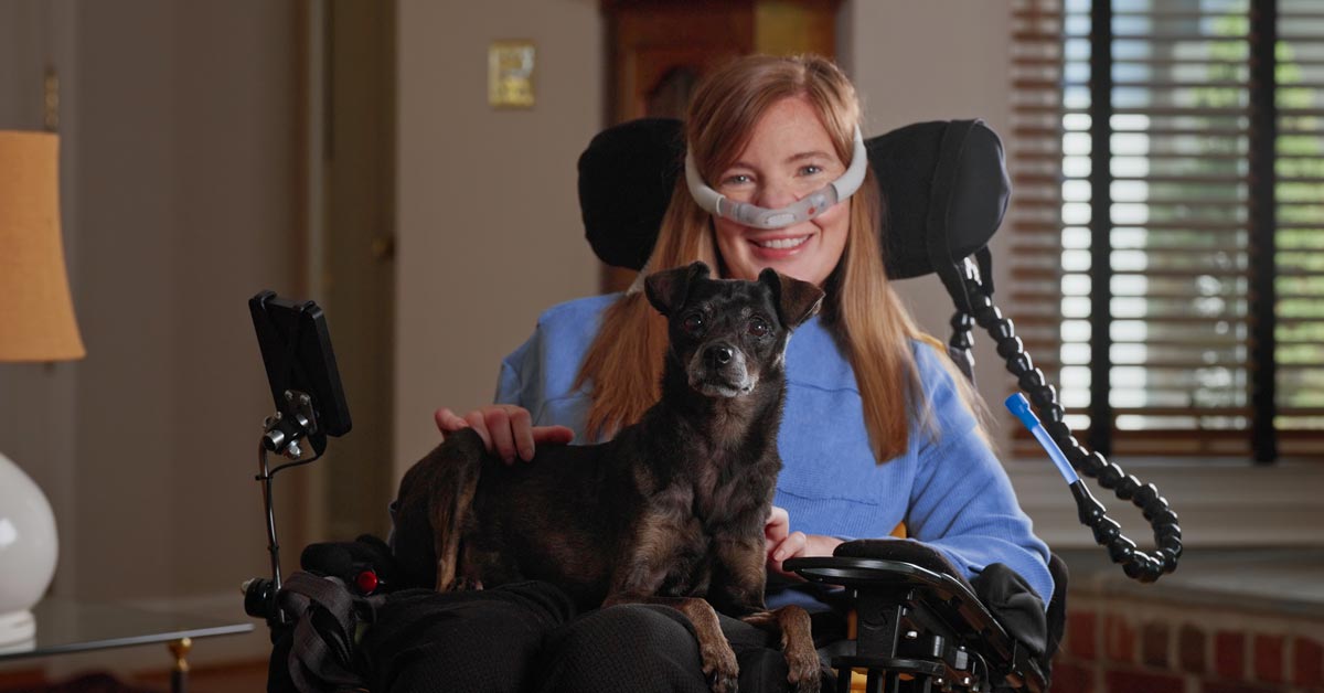 A woman with ALS, Brooke Eby, sits in a wheelchair with an oxygen tube. She holds a small, black dog on her lap