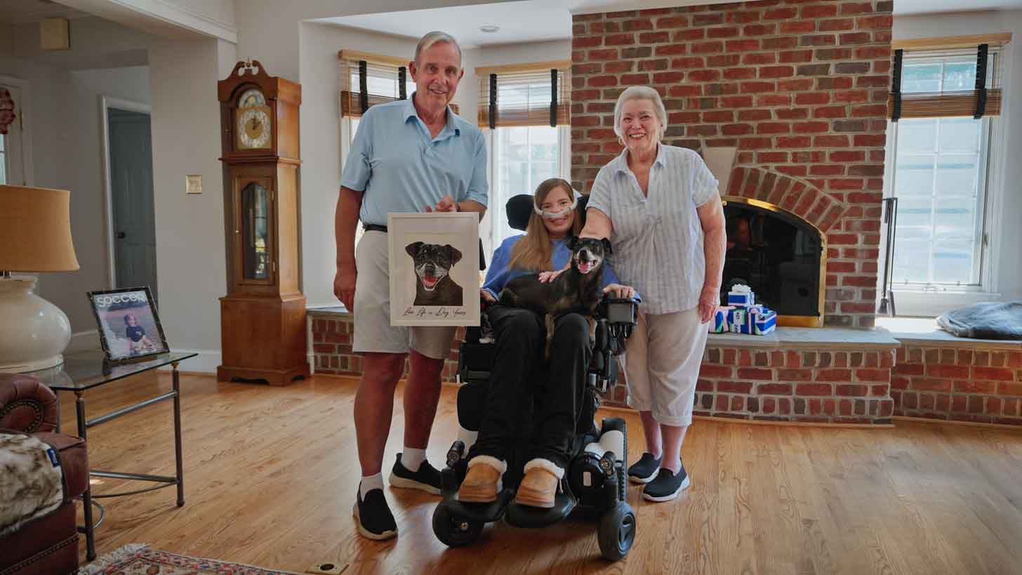 A woman with ALS, Brooke Eby, sits in a wheelchair with an oxygen tube. She holds a small, black dog on her lap. Two older adults, her parents, stand beside her, smiling.
