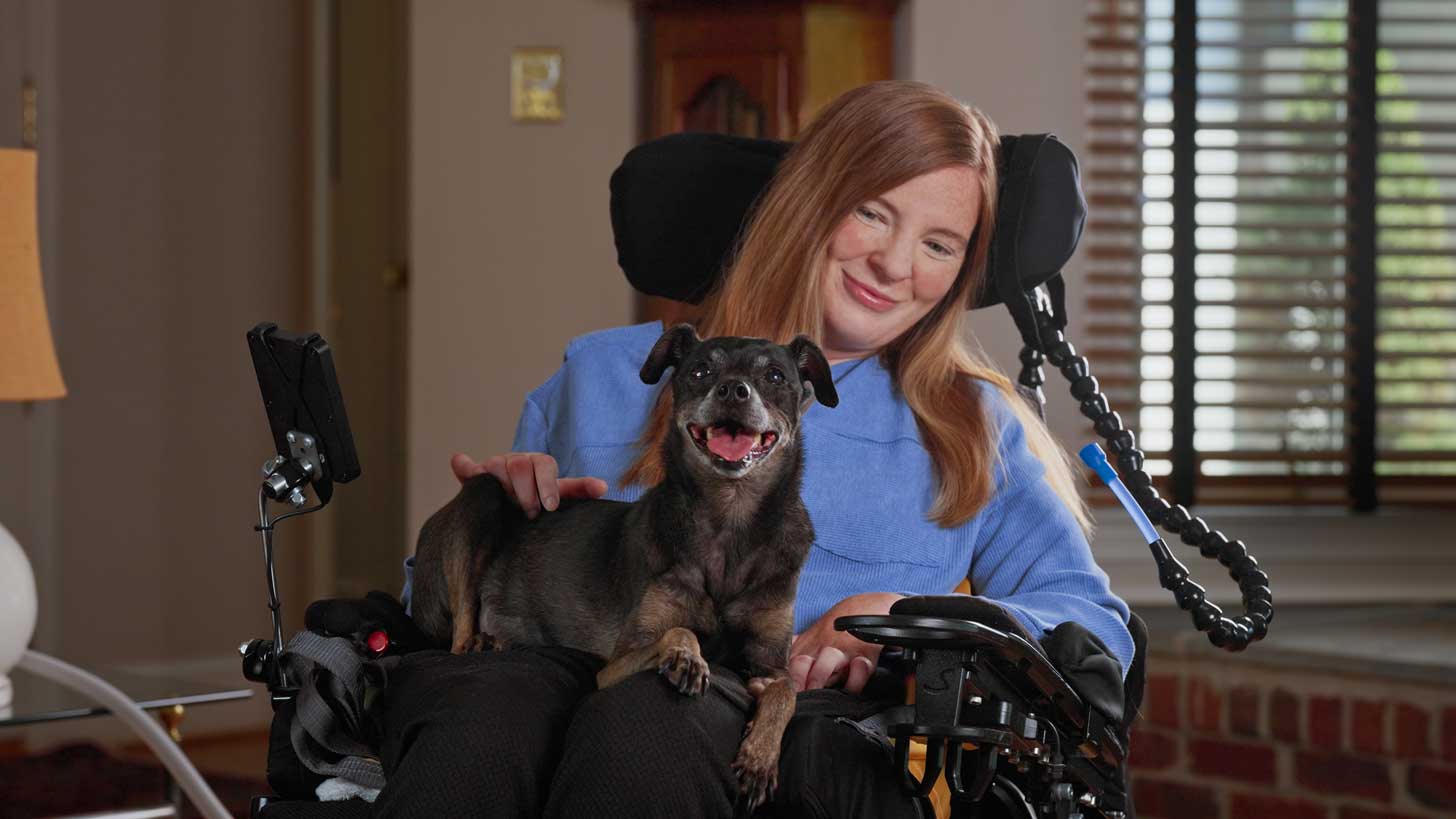 A woman with ALS, Brooke Eby, sits in a wheelchair. She holds a small, black dog on her lap