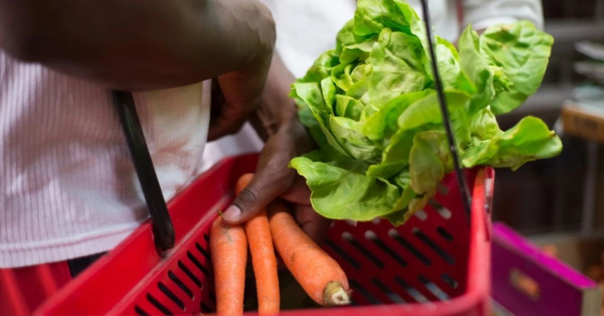 A close-up image of a Black man putting carrots and lettuce into a grocery cart.