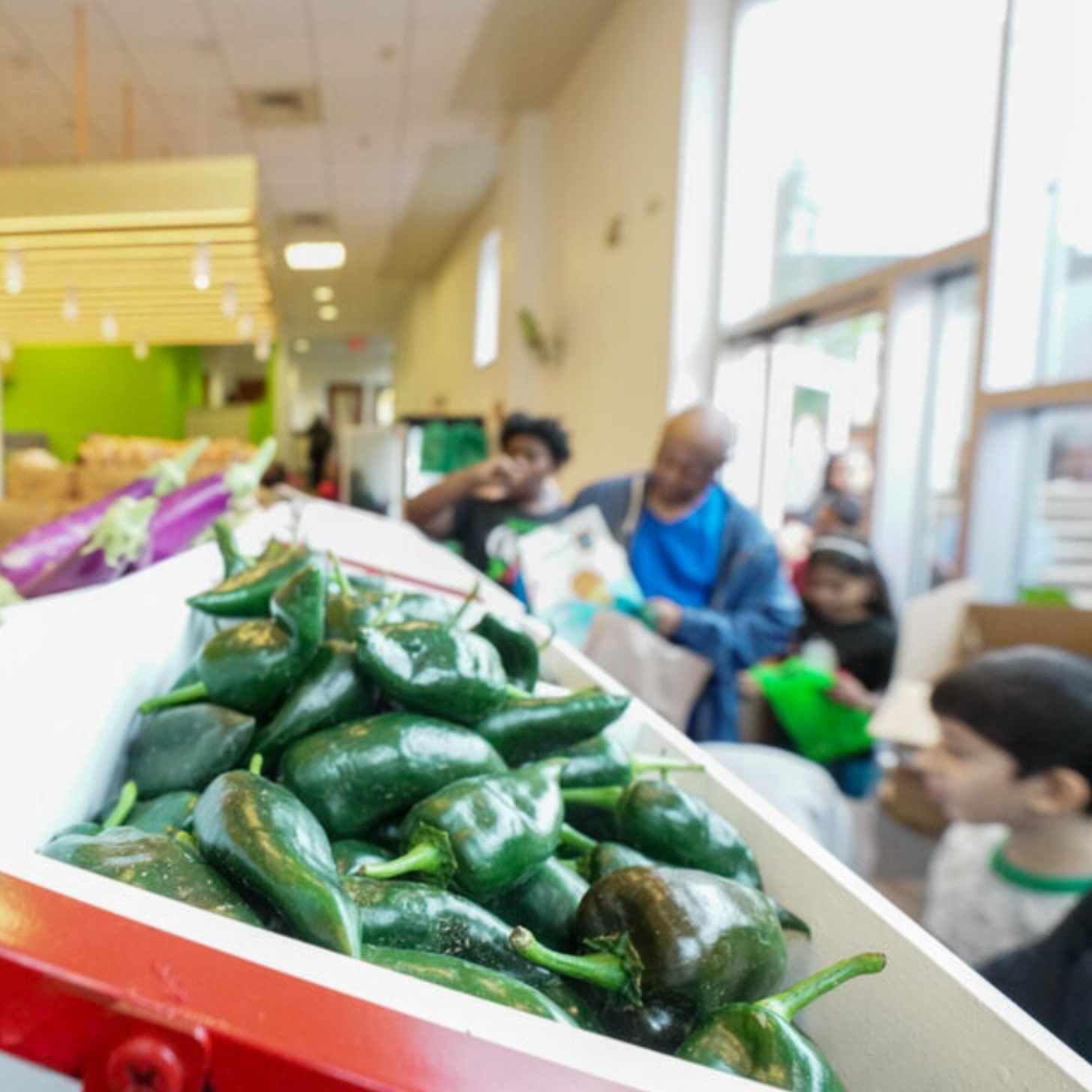 A crowd of shoppers walk through a grocery store. The image is focused on a bin of fresh green peppers.