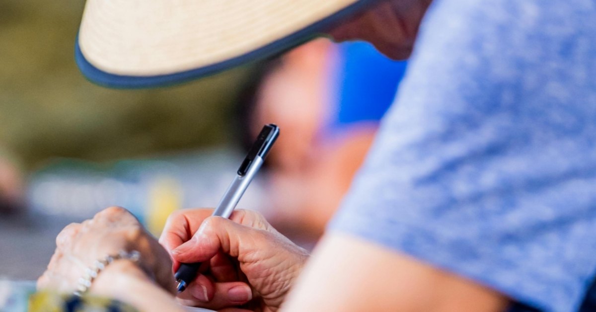 An older woman is crouched by a table writing something down as her hat dips into frame