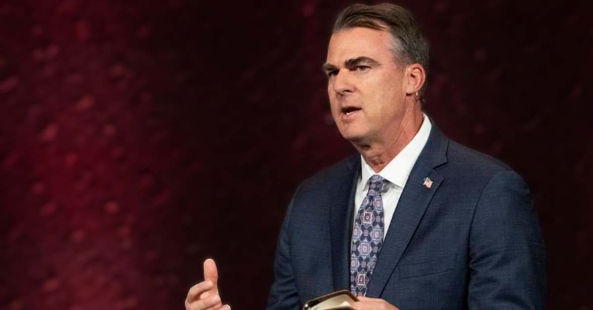 J. Kevin Stitt, a white man with brown gray hair addresses a room of people in front of a dark red background.