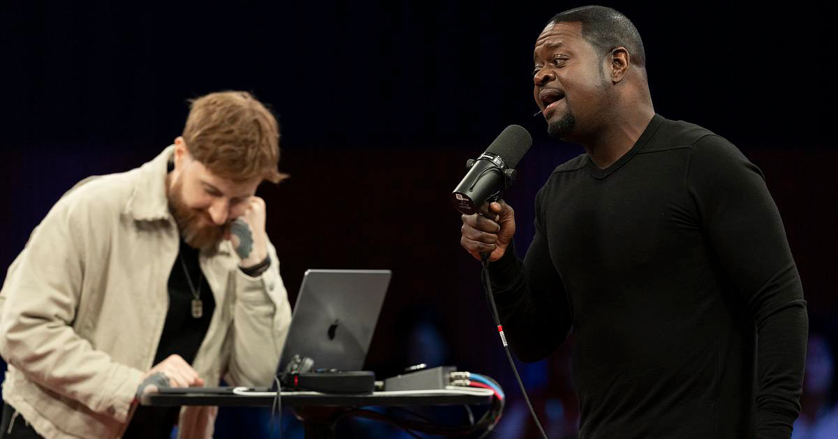 Jason Boyd — Poo Bear — holds a microphone to his mouth, singing, while an accompanist operates a laptop
