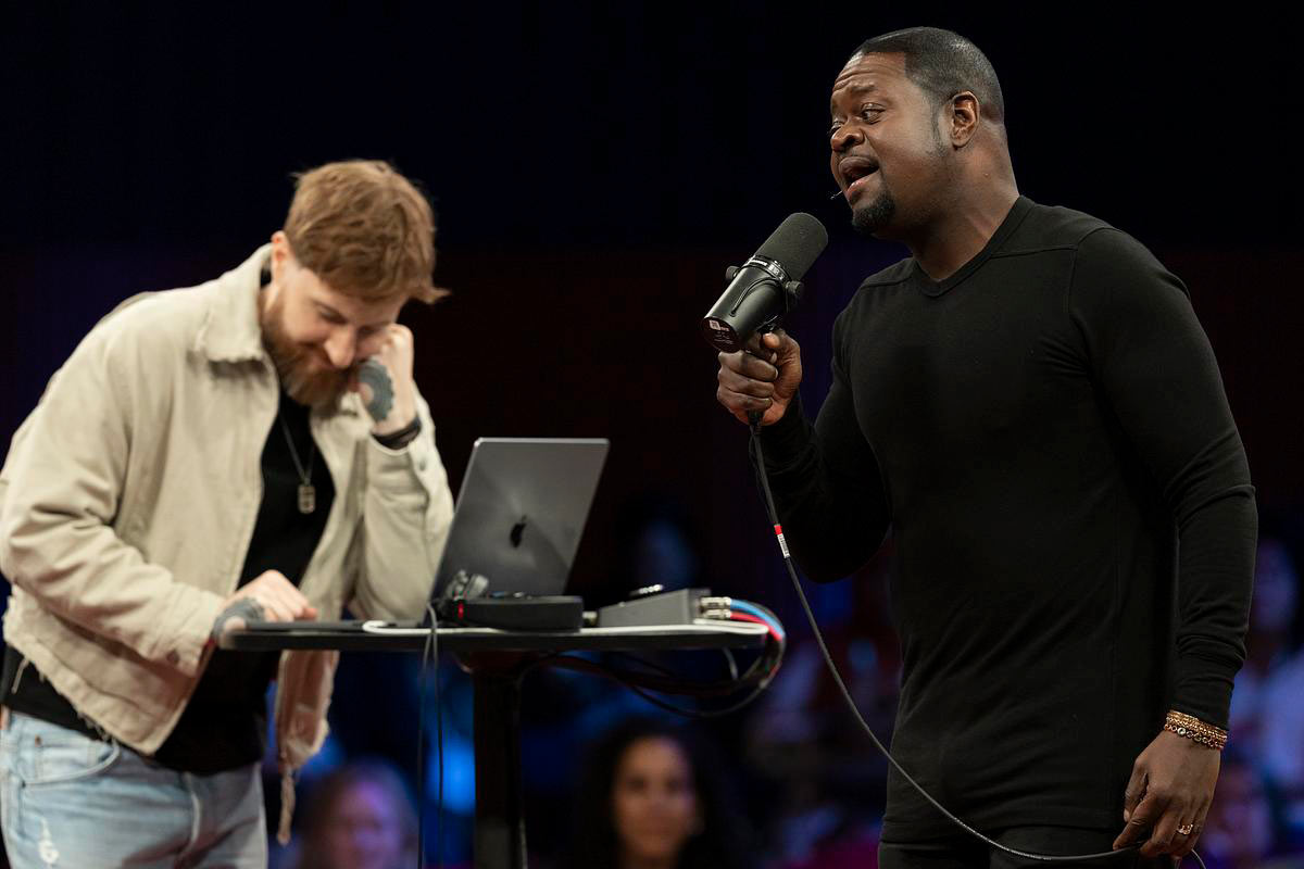Jason Boyd — Poo Bear — holds a microphone to his mouth, singing, while an accompanist operates a laptop