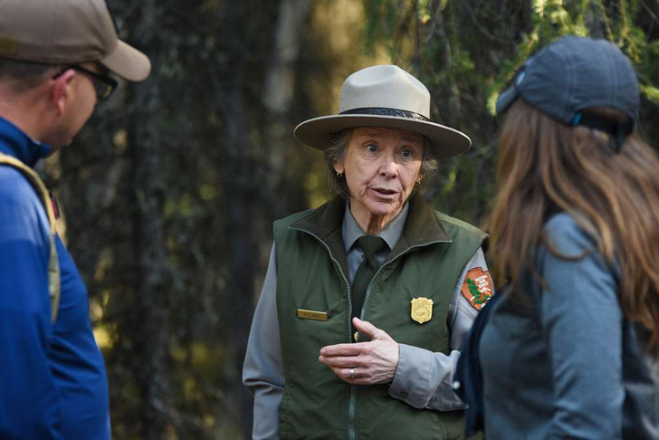 An elderly female national park ranger speaks to two visitors