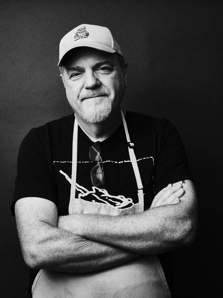 A black and white photo of a man in a baseball cap and apron, with his arms crossed. He is Chris Hyde, the founder of Souper Sunday
