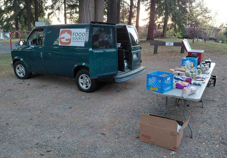 A green van has a logo for the Food Source Location and is open to tables of groceries in a park in Olympia, Washington