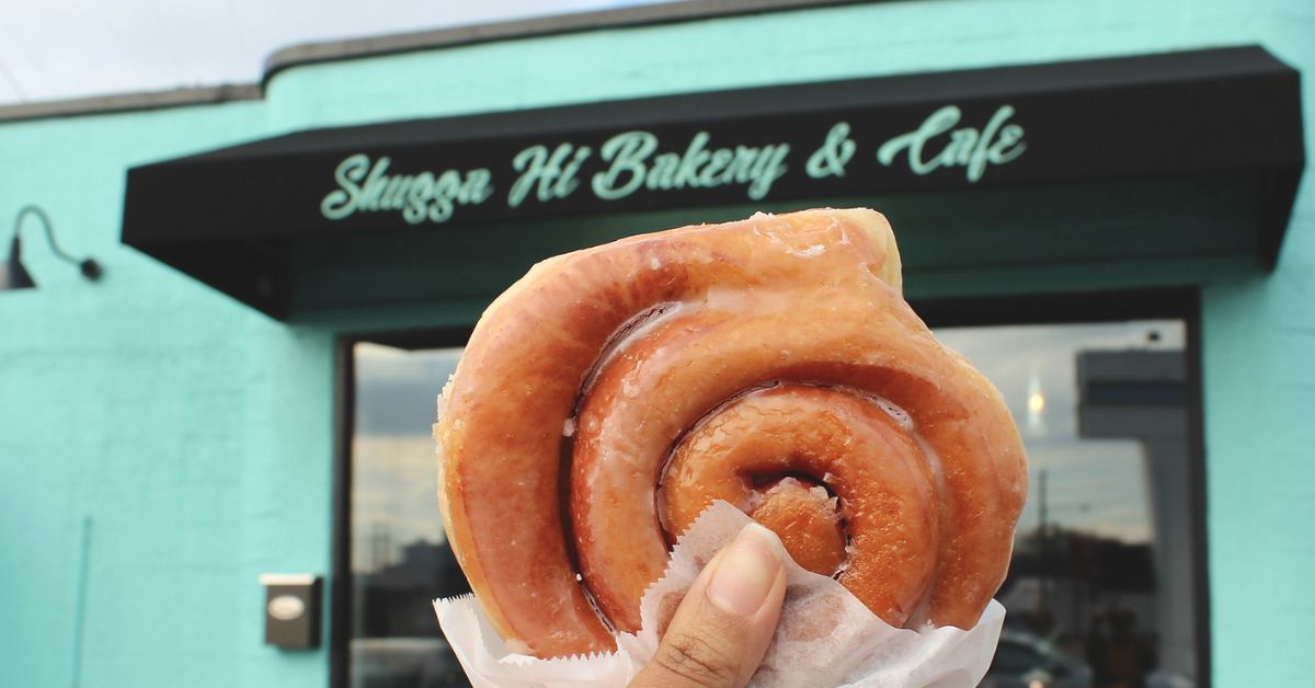 A person holds a cinnamon roll up in front of a teal bakery front that says: Shugga Hi Bakery & Cafe 