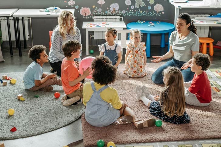 Two child care workers sit on a carpet, talking to a diverse group of children