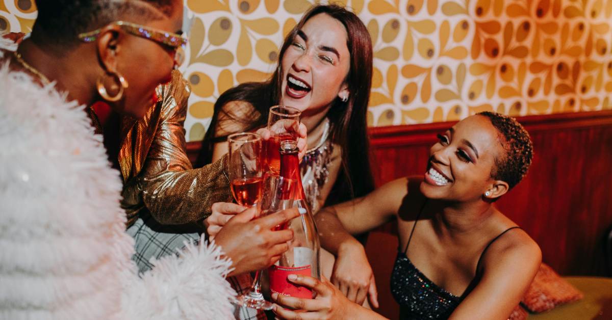 A group of three women of varying ethnicities laugh as they hang out at a bar, pouring drinks