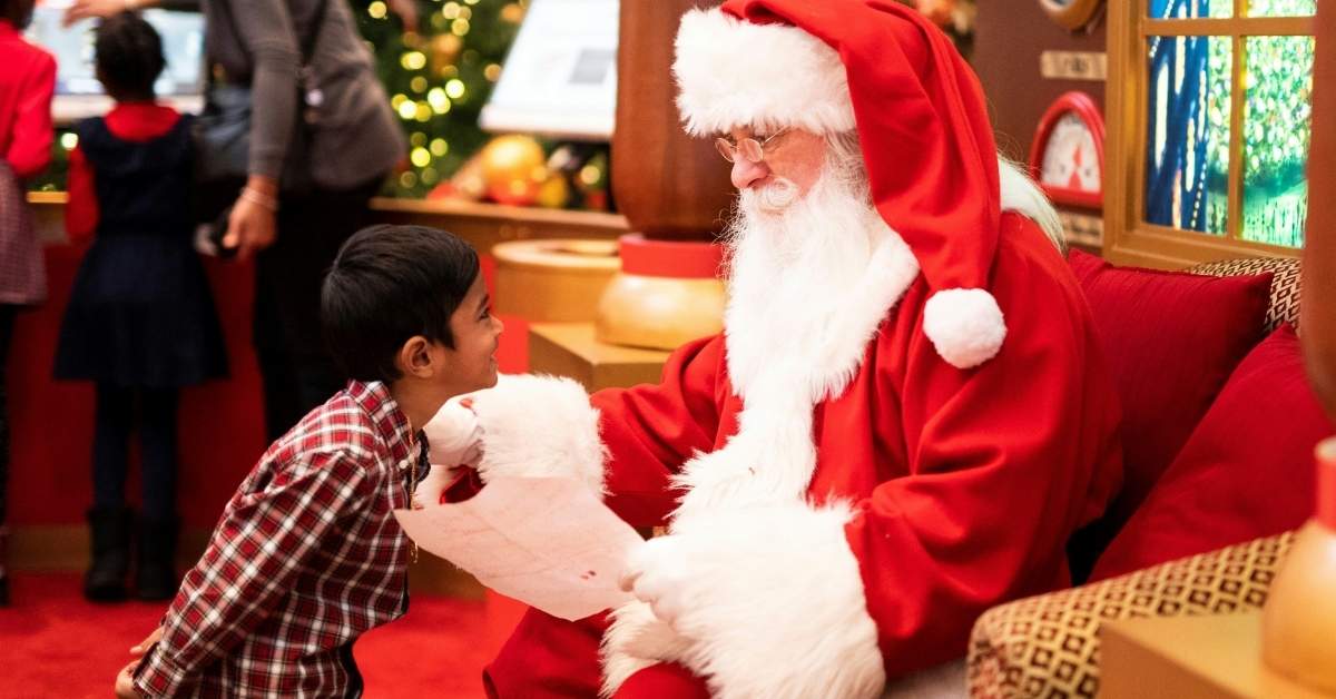 A boy stands in front of a man wearing a Santa Claus costume
