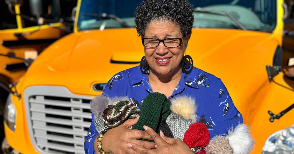 A bus driver, Tina Hutcherson, holds a bundle of knitted beanies in her arms while standing and smiling in front of a school bus