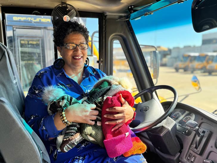 A bus driver, Tina Hutcherson, sits at her post in a school bus, holding an array of knitted beanies in her arms.