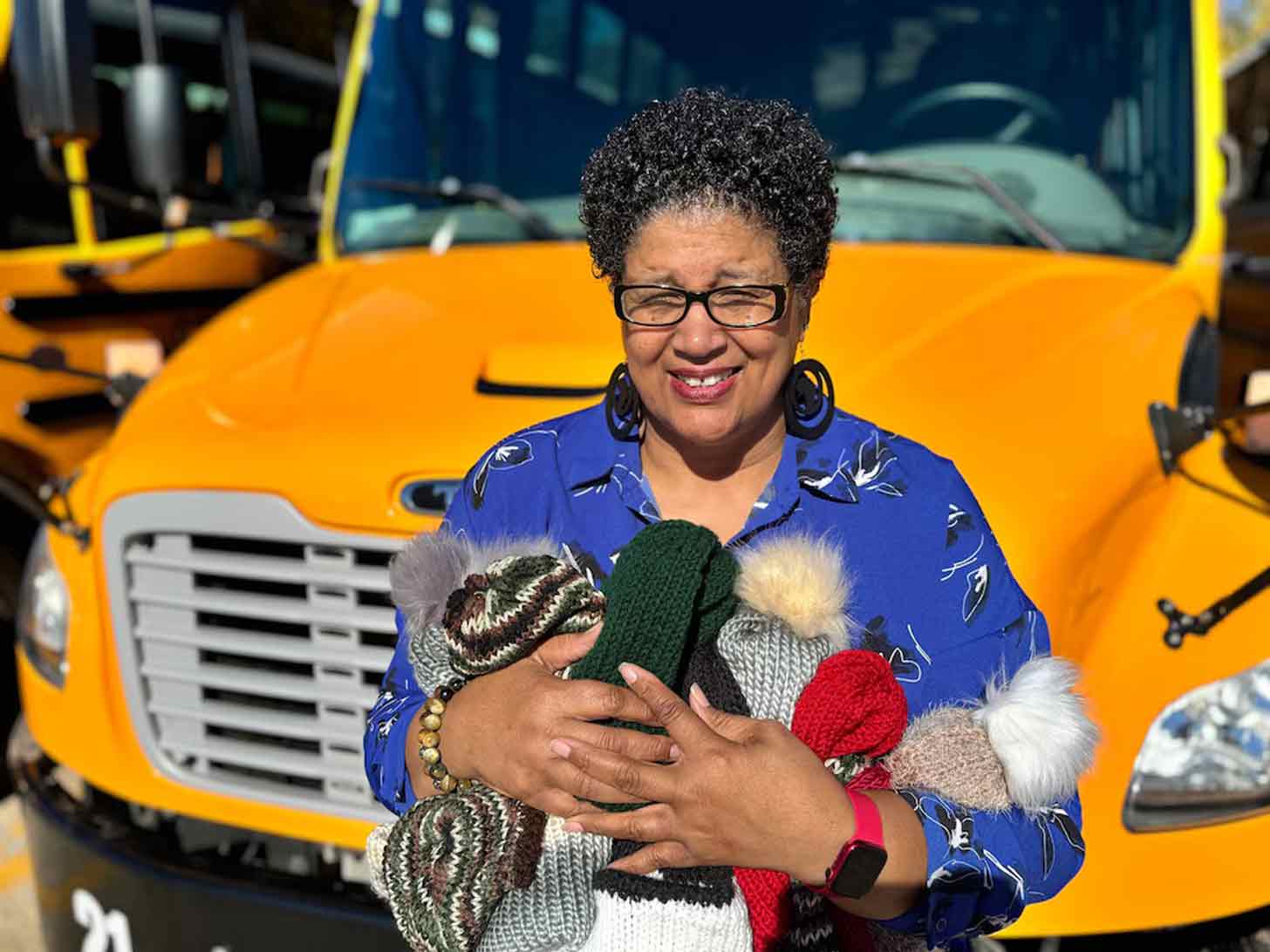 A bus driver, Tina Hutcherson, holds a bundle of knitted beanies in her arms while standing and smiling in front of a school bus