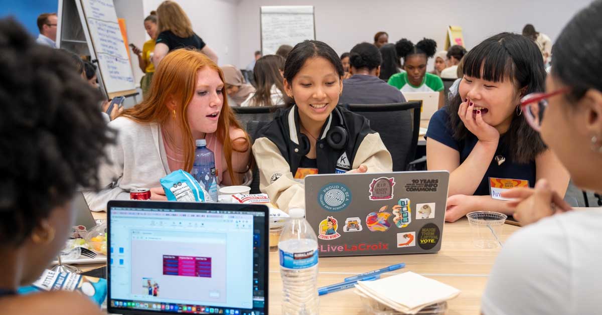 A group of young girls gather around laptops at a Girls Who Code club, laughing and smiling