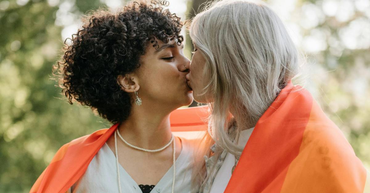 A Black woman with short hair kisses a white woman with blonde hair with a pride flag wrapped around their shoulders. 