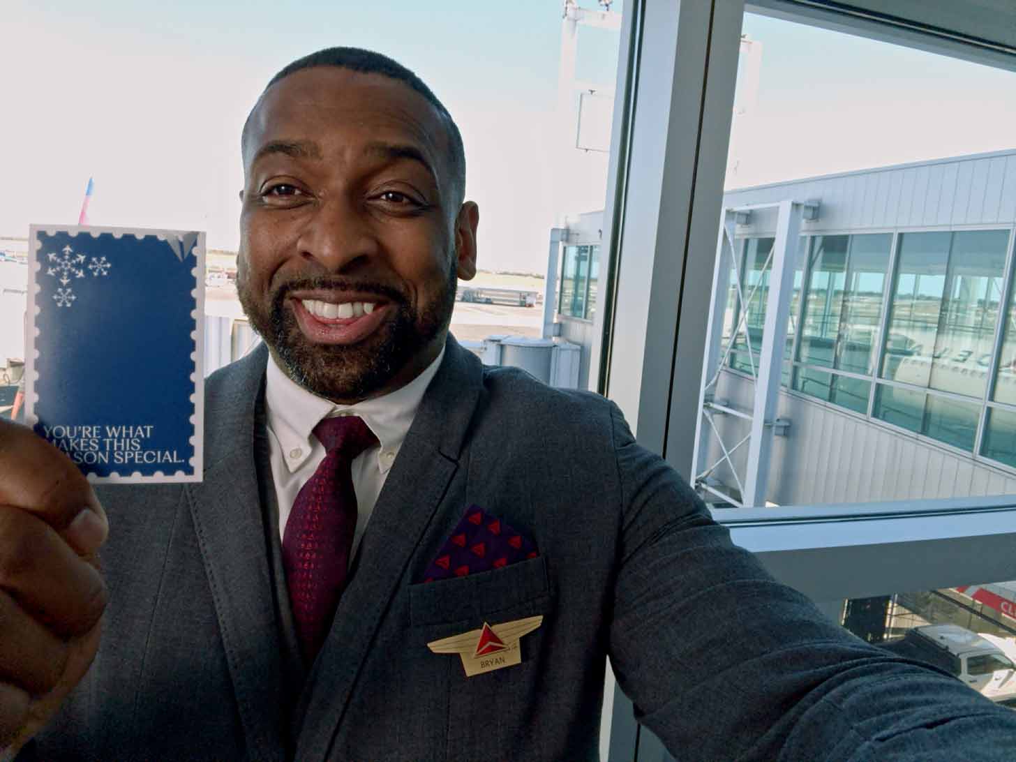 A Black man and Delta airlines employee in a gray suit jacket takes a selfie in an airport, holding up a gift card