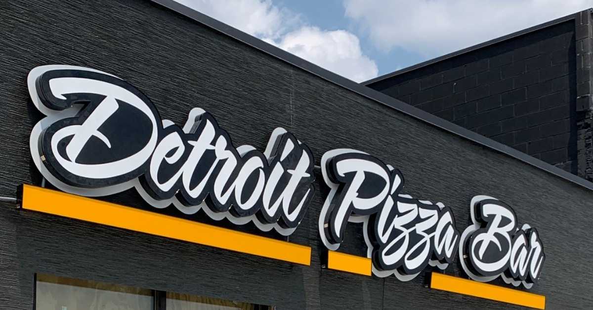 The exterior of a restaurant called Detroit Pizza Bar. It has a cursive style font in white lettering on a black tile roof.