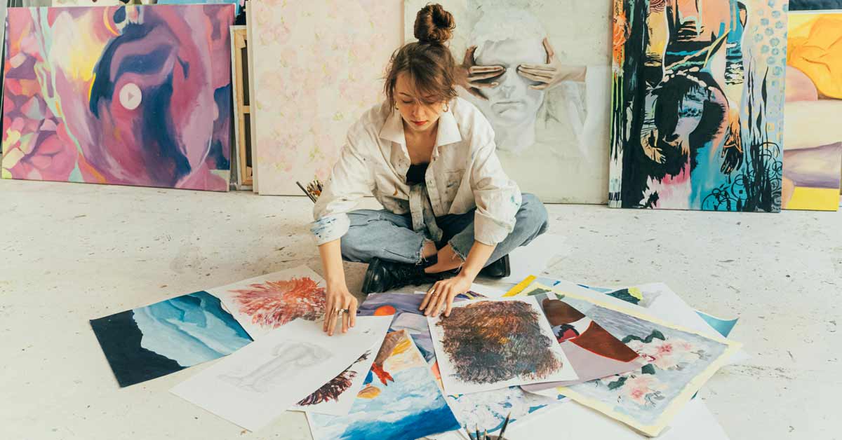 A woman sits cross-legged on the floor of an art studio, studying a handful of paintings