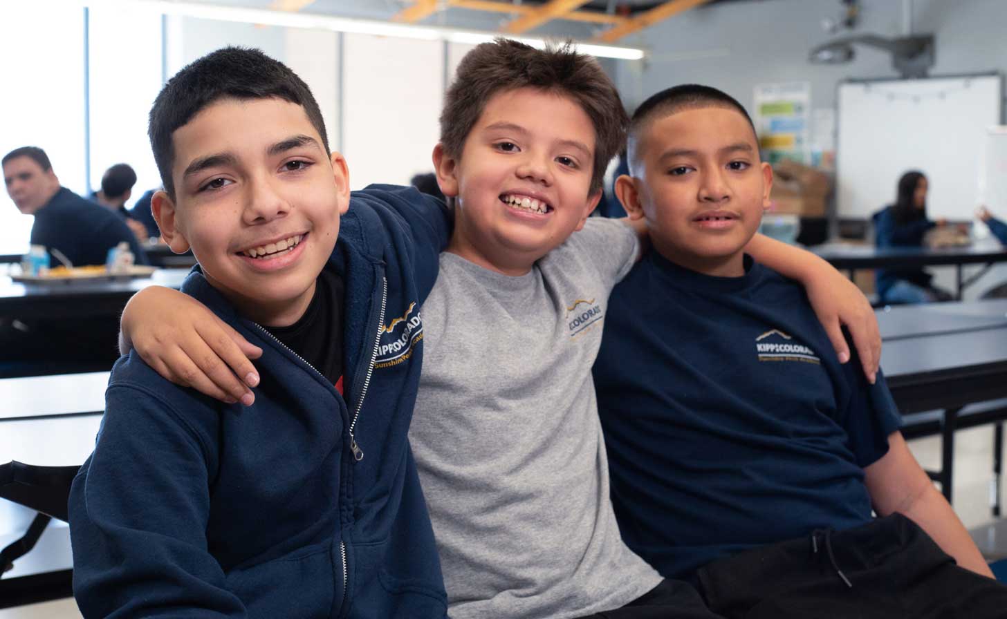 Three young boys with their arms around each other in a school cafeteria