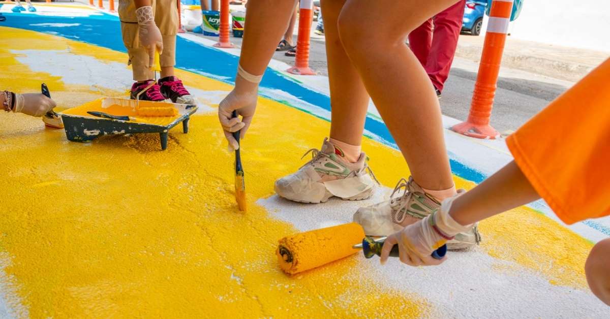 Children (photographed from the knees down) paint a sidewalk yellow