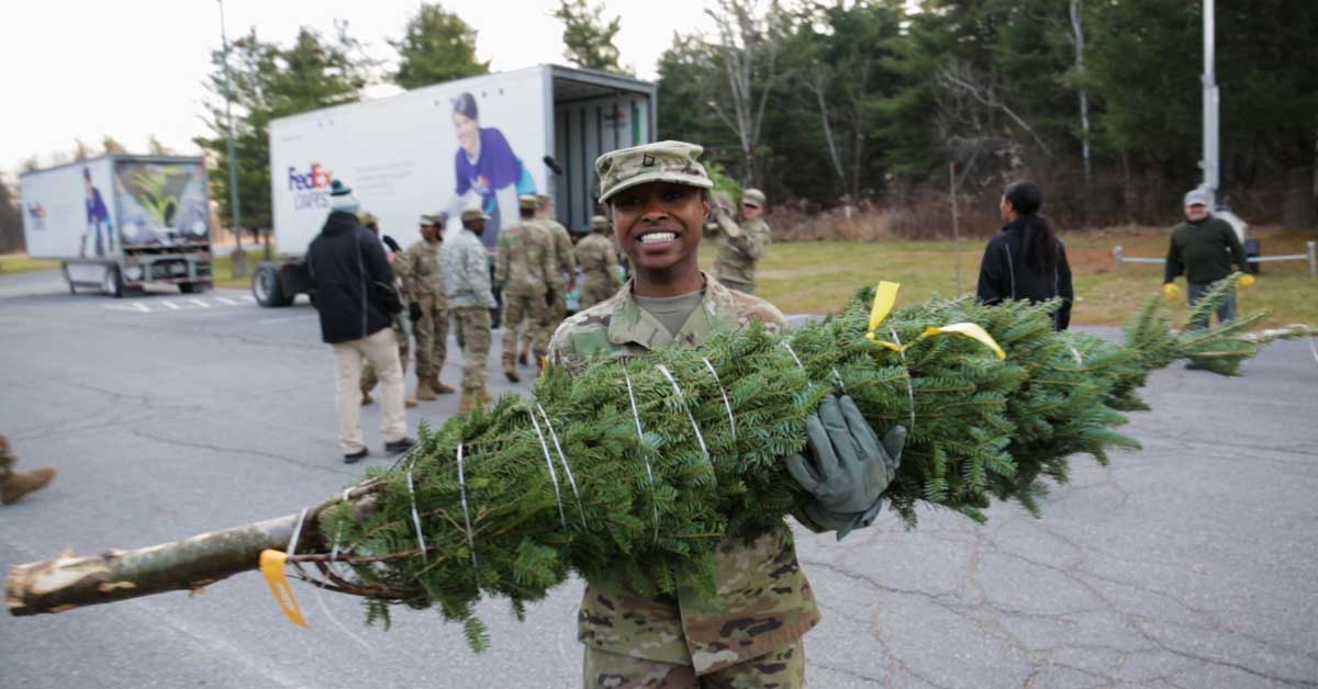 A Black woman wears military fatigues and holds a Christmas tree in her arms in front of a FedEx truck