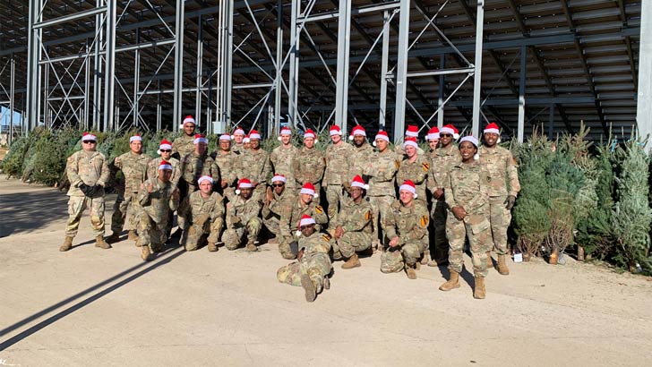 A group of people in military fatigues and Santa hats stand in front of a collection of Christmas trees
