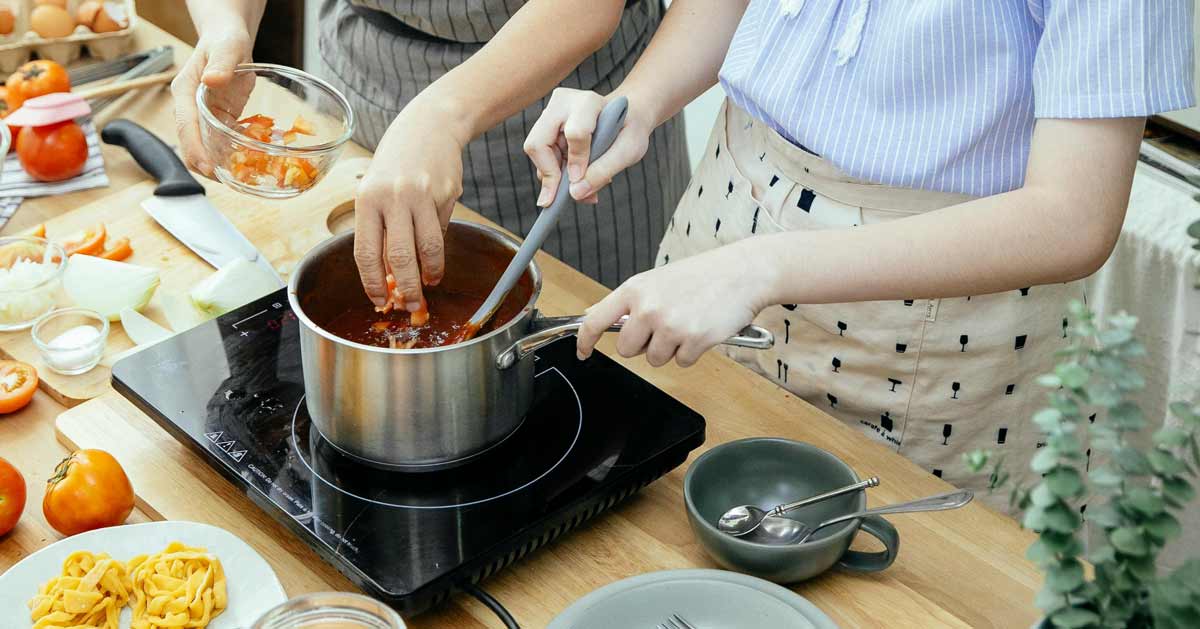 A person stirs a pot while another puts ingredients in