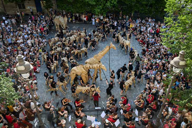 A crowd of people gather around a herd of cardboard animal puppets in Paris